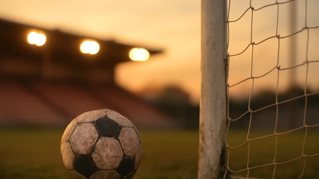 Balón junto a la red en portería, gradas vacías y focos encendidos.